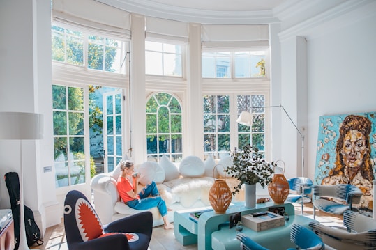 A bright, modern living room with large windows letting in abundant natural light. A person is sitting on a white sofa, using a smartphone. The room features eclectic decor, including a unique black chair with a shark-like design, a light blue coffee table with plants and decorative objects, and a large, colorful portrait painting leaning against the wall. The walls are white, and the overall atmosphere is airy and inviting.