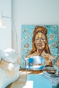 A modern, stylish living space featuring a large, textured painting of a face with earthy tones against a turquoise background. The room includes a sleek floor lamp, a cozy white chair with a fluffy throw, an elegant blue chair, and a stack of books or magazines on a blue table. Natural light streams in, creating a bright and inviting atmosphere.