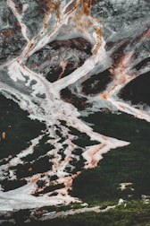 A dramatic landscape featuring rugged, steep mountains with striking white and orange mineral deposits. Dark green forests cover the lower parts of the scene, contrasting sharply with the rocky terrain. The patterns formed by the mineral deposits create a visually intriguing texture.