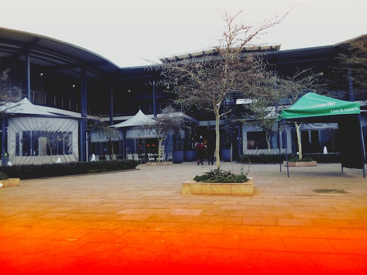 An outdoor courtyard area featuring several leafless trees surrounded by modern architectural buildings. The setup includes outdoor seating under covered awnings and a tent with 'Live it. Love it.' written on it. The foreground shows tiled paving with a gradient effect in shades of red and orange.