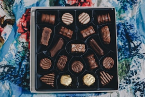 An open box contains a variety of chocolates, each with different shapes and patterns, resting on a bed with a colorful, abstract-patterned fabric covering.