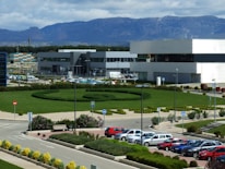 An industrial or office complex with modern buildings surrounded by a well-maintained garden. There is a large, circular hedge with paths running through the landscaped area. Multiple parked cars are visible in the foreground, and a backdrop of mountains under a partly cloudy sky enhances the scenery.