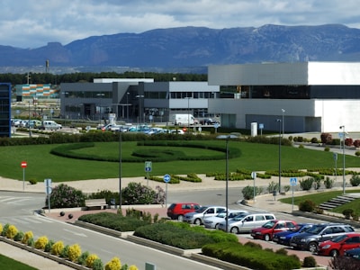 An industrial or office complex with modern buildings surrounded by a well-maintained garden. There is a large, circular hedge with paths running through the landscaped area. Multiple parked cars are visible in the foreground, and a backdrop of mountains under a partly cloudy sky enhances the scenery.