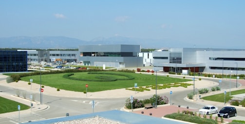 A modern industrial or office complex with sleek, geometric buildings in a gray and white color palette. The area features a large roundabout with manicured grass and small shrubs in the center. Several parked cars are visible along the roads, and there are clear blue skies in the background with distant mountain ranges on the horizon.