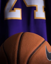 A close-up view of a textured basketball positioned in front of a purple sports jersey with the number 24 in white and yellow.