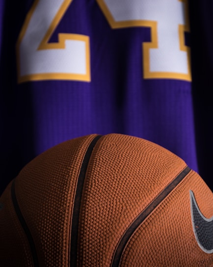 A close-up view of a textured basketball positioned in front of a purple sports jersey with the number 24 in white and yellow.