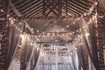 An interior view of a rustic barn adorned with hanging string lights, which provide a warm glow. Heavy, translucent drapes hang down the sides, creating a cozy and elegant atmosphere in an otherwise simple wooden structure. The ceiling's wooden beams form an intricate pattern, leading the eye down the length of the space.