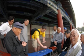 A group of elderly men, along with a younger individual wearing a yellow shirt, are gathered around a table outside a traditional building with ornate, painted eaves. They appear to be engaged in a game or activity involving tiles on the table. The scene is informal and social.