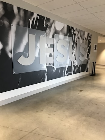 A large mural on a wall with the word 'JESUS' prominently displayed in bold letters. The background appears to feature raised hands and arms, possibly in a gesture of praise or worship. The setting seems to be a modern indoor hallway or lobby with polished concrete floors and a white ceiling.