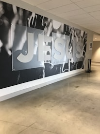 A large mural on a wall with the word 'JESUS' prominently displayed in bold letters. The background appears to feature raised hands and arms, possibly in a gesture of praise or worship. The setting seems to be a modern indoor hallway or lobby with polished concrete floors and a white ceiling.