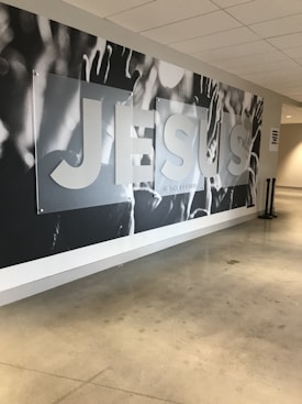 A large mural on a wall with the word 'JESUS' prominently displayed in bold letters. The background appears to feature raised hands and arms, possibly in a gesture of praise or worship. The setting seems to be a modern indoor hallway or lobby with polished concrete floors and a white ceiling.