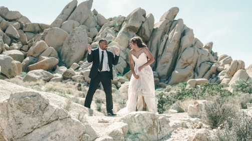 A couple dressed in formal attire is standing in a rocky desert landscape. The man in a black suit and white shirt seems energetic, while the woman in a flowing white dress looks joyful as they interact with each other. The background features large boulders and sparse desert vegetation under a clear blue sky.