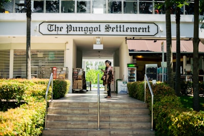 The image features the entrance to a building labeled 'The Punggol Settlement.' The scene includes lush green bushes lining the stairs leading up to the entrance. A person is seen standing on the steps, facing away from the camera. Various retail elements, such as vending machines and market stands, are visible in the background under the awning.