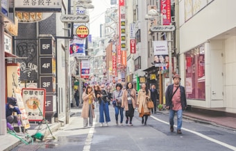 A bustling street scene in Tokyo with people walking along a narrow road lined with various shops and signs in Japanese. Vibrant advertisements and colorful shop fronts create a lively atmosphere. The individuals appear engaged, with some using their phones and others conversing. The street is clean, and the urban environment is well-maintained.