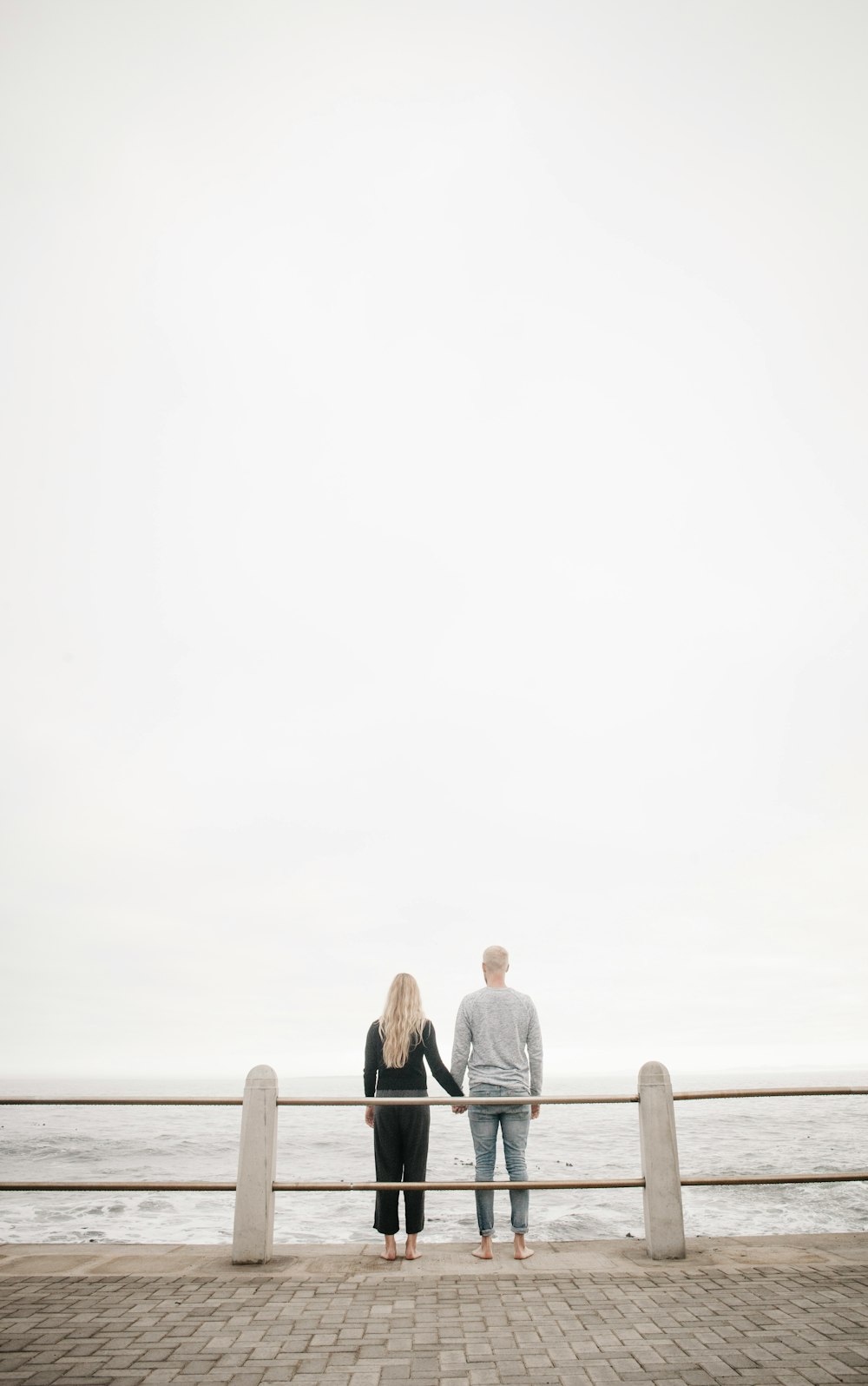 couple leaning on guard rail while holdings hands facing ocean at daytime