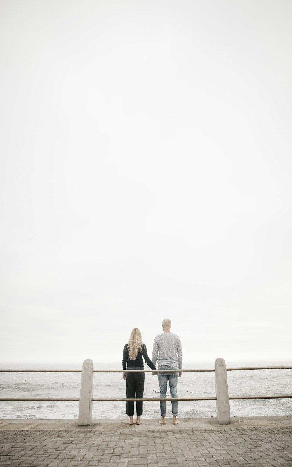 Paar lehnt sich an Leitplanke, während es tagsüber Händchen mit Blick auf den Ozean hält