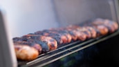 Several sausages are lined up on a grill, slightly charred and emitting smoke. The focus is on the sausages with a soft, blurred background, suggesting a close-up perspective. The sausages appear to be cooking outdoors.