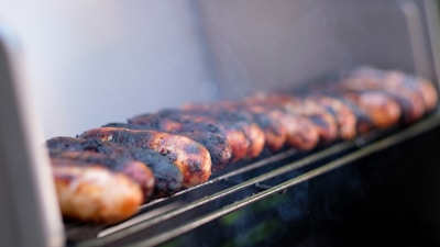 Several sausages are lined up on a grill, slightly charred and emitting smoke. The focus is on the sausages with a soft, blurred background, suggesting a close-up perspective. The sausages appear to be cooking outdoors.