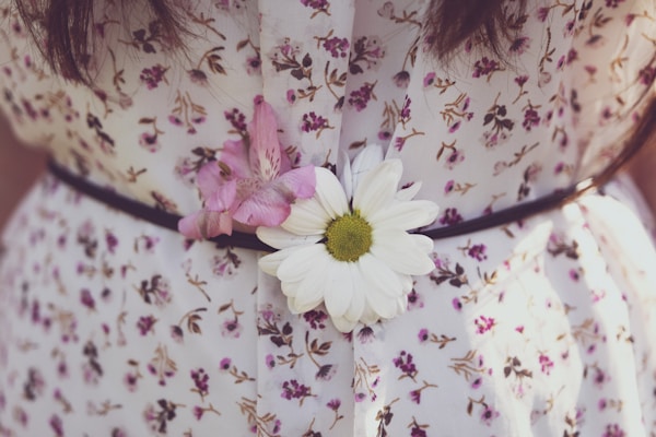 A close-up of a floral patterned dress with a wide black belt. Two flowers, a white daisy and a pink flower, are tucked into the belt, adding a natural accent to the outfit.