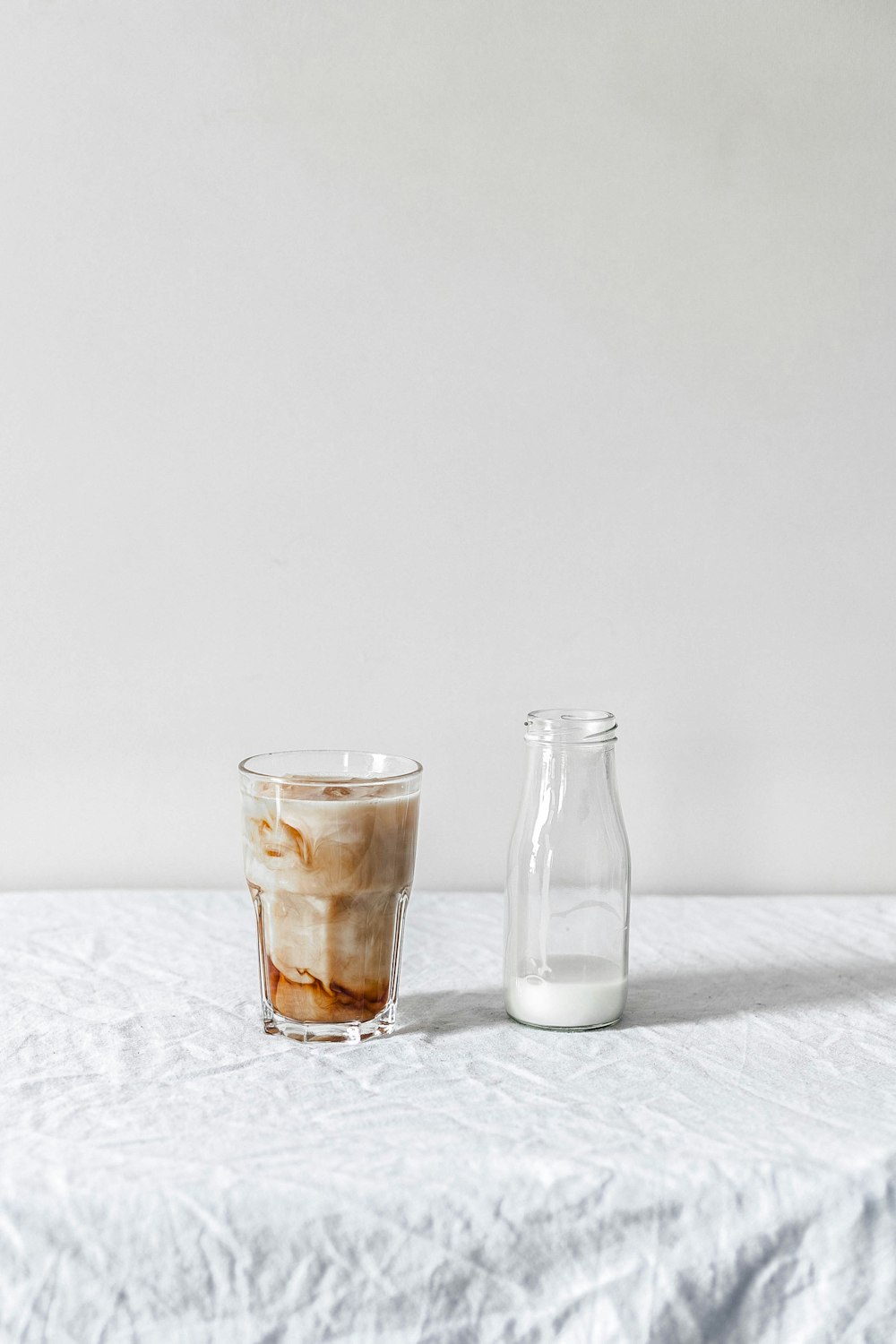 botella de vidrio transparente llena de bebida blanca junto a un vaso de pinta transparente lleno de líquido marrón