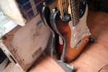 A sunburst electric guitar leaning against a white, rugged flight case. The guitar has a typical double-cutaway design with a white pickguard and several control knobs. The flooring appears to be wooden.
