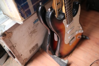 A sunburst electric guitar leaning against a white, rugged flight case. The guitar has a typical double-cutaway design with a white pickguard and several control knobs. The flooring appears to be wooden.