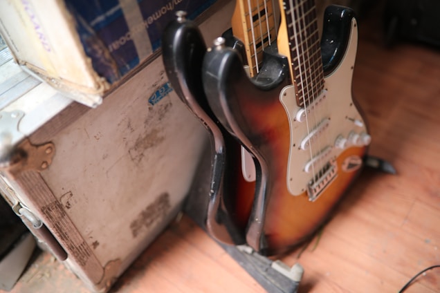 A sunburst electric guitar leaning against a white, rugged flight case. The guitar has a typical double-cutaway design with a white pickguard and several control knobs. The flooring appears to be wooden.