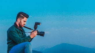 A person with dark hair is wearing a green shirt and holding a camera with a large lens and a flash attachment. The individual is sitting outdoors, seemingly focused on taking a photo. The sky is clear with a blue hue, and there are subtle mountains visible in the distance.