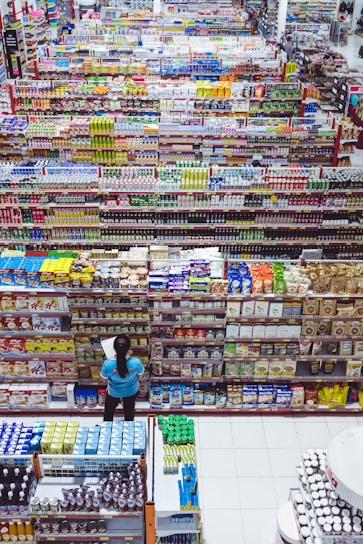 A large supermarket aisle displays a variety of colorful packaged goods arranged neatly on shelves. A person in a blue shirt stands in front of the shelves, holding a list or paper, likely checking items or inventory. The view from above shows an extensive selection of products, indicating a wide variety of options available to shoppers.
