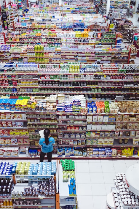 A large supermarket aisle displays a variety of colorful packaged goods arranged neatly on shelves. A person in a blue shirt stands in front of the shelves, holding a list or paper, likely checking items or inventory. The view from above shows an extensive selection of products, indicating a wide variety of options available to shoppers.