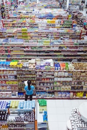 A large supermarket aisle displays a variety of colorful packaged goods arranged neatly on shelves. A person in a blue shirt stands in front of the shelves, holding a list or paper, likely checking items or inventory. The view from above shows an extensive selection of products, indicating a wide variety of options available to shoppers.