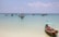 Small fishing boats are floating on a calm turquoise sea, with a clear blue sky above. A single boat with a turquoise canopy is anchored close to the shore, while several other boats are scattered in the distance.