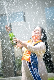 A person in a lace dress with graduation stoles is joyfully spraying champagne. The spray creates a lively waterfall of bubbles and droplets around them. The setting appears outdoors with a blurred background highlighting the celebratory moment.