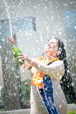 A person in a lace dress with graduation stoles is joyfully spraying champagne. The spray creates a lively waterfall of bubbles and droplets around them. The setting appears outdoors with a blurred background highlighting the celebratory moment.