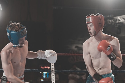 Two boxers are engaged in a match inside a boxing ring. Both are wearing protective headgear and boxing gloves, one with red gloves and headgear, the other with blue. The boxer in red is delivering a punch, while the boxer in blue is on the defensive. The background is dimly lit, giving focus to the competitors.