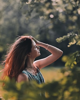 A person with long, brown hair stands in a lush, green setting, gently touching their head and looking serene. Sunlight filters through the leaves, casting a warm glow and creating a peaceful atmosphere.