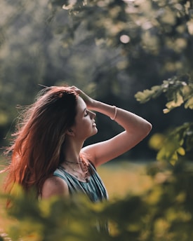 A person with long, brown hair stands in a lush, green setting, gently touching their head and looking serene. Sunlight filters through the leaves, casting a warm glow and creating a peaceful atmosphere.