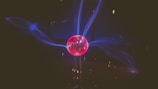 A glowing spherical object emits blue electrical arcs against a dark background. The center of the sphere is bright pink, and the electric arcs create a dynamic, swirling pattern extending outward from the sphere.
