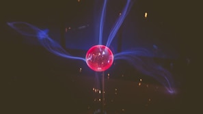 A glowing spherical object emits blue electrical arcs against a dark background. The center of the sphere is bright pink, and the electric arcs create a dynamic, swirling pattern extending outward from the sphere.