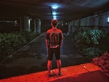 A person stands under a dimly lit underpass, facing away, wearing a dark sports jersey with the number 10 and the name 'Sakuragi' on the back. The area is illuminated by overhead lights, casting a bluish tone, and is surrounded by green bushes. The ground has a red hue near the bottom of the image, adding a contrasting color.