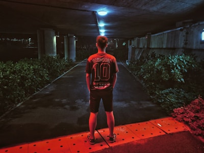 A person stands under a dimly lit underpass, facing away, wearing a dark sports jersey with the number 10 and the name 'Sakuragi' on the back. The area is illuminated by overhead lights, casting a bluish tone, and is surrounded by green bushes. The ground has a red hue near the bottom of the image, adding a contrasting color.