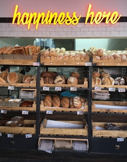 A variety of freshly baked bread arranged on wooden shelves in a bakery setting. A neon sign above reads 'happiness here.' The shelves are filled with different types of bread, including baguettes, round loaves, and seeded bread.