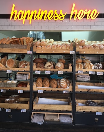 A variety of freshly baked bread arranged on wooden shelves in a bakery setting. A neon sign above reads 'happiness here.' The shelves are filled with different types of bread, including baguettes, round loaves, and seeded bread.