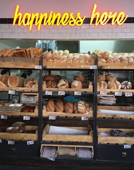 A variety of freshly baked bread arranged on wooden shelves in a bakery setting. A neon sign above reads 'happiness here.' The shelves are filled with different types of bread, including baguettes, round loaves, and seeded bread.