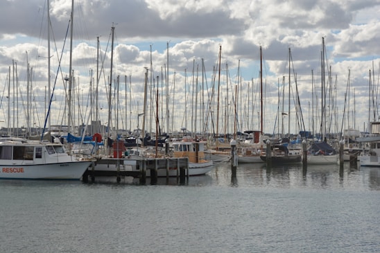 A harbor filled with numerous sailboats and yachts moored at docks. Tall masts reach up towards a sky filled with fluffy clouds. The water is calm, reflecting the array of vessels. One boat has 'Rescue' written on its side.