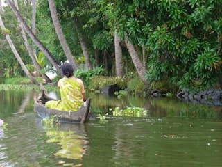 A woman in a green sari is paddling a small wooden canoe on a peaceful river surrounded by lush greenery and palm trees. There are water plants floating on the surface of the water.