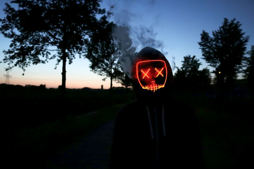 A model wearing a tattered black hoodie with eerie glowing red eyes printed on the back, standing in a foggy graveyard at dusk.
