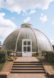 A large, dome-shaped greenhouse with a glass structure dominates the scene. The front entrance has a white wooden door with glass panels. Stone steps lead up to the entrance, flanked by potted plants. The sky above is clear with a few fluffy clouds, giving a bright and airy atmosphere.