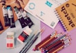 A variety of art supplies are spread out, including tubes of paint, jars of medium, colored pencils, a sketchbook, and sheets of paper. There are several artist paint brushes alongside pencils of different colors arranged neatly, with some paint tubes and medium bottles placed nearby.