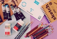 A variety of art supplies are spread out, including tubes of paint, jars of medium, colored pencils, a sketchbook, and sheets of paper. There are several artist paint brushes alongside pencils of different colors arranged neatly, with some paint tubes and medium bottles placed nearby.